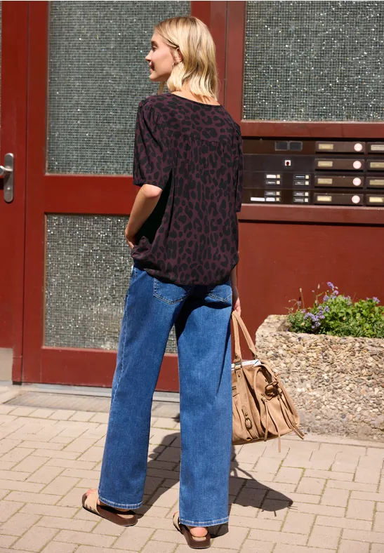 Person von hinten vor einer Tür, in weiten blauen Jeans, schwarzem T-Shirt mit rotem Muster, Sandalen und Tasche.