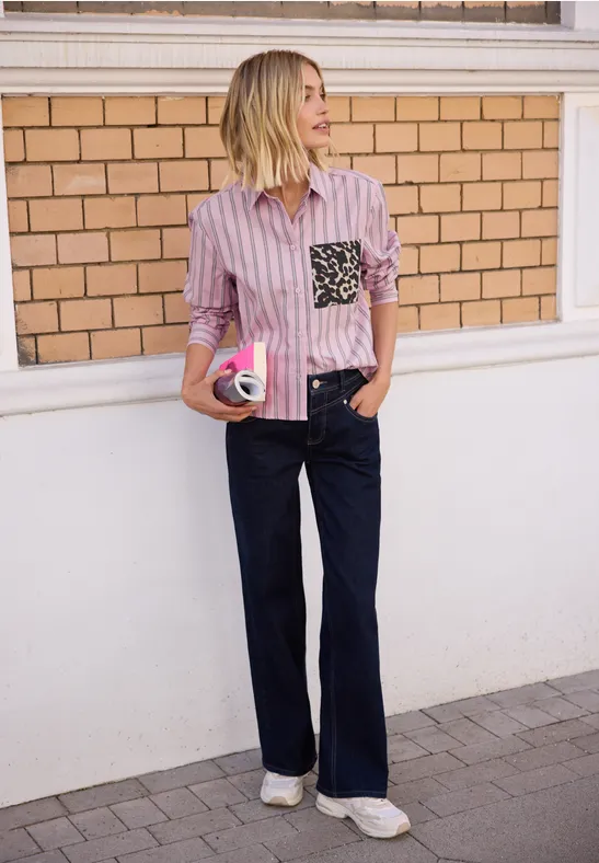 Femme en chemise rose à rayures avec poche noire, jean large et baskets blanches, tenant un café devant un mur de briques.