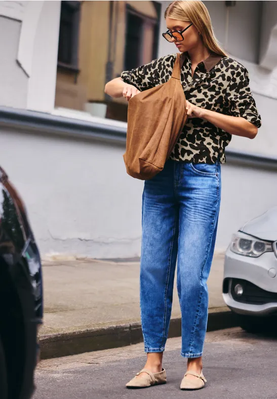 Frau in Jeans und Leopardenmuster-Bluse mit Tasche auf Gehweg zwischen parkenden Autos