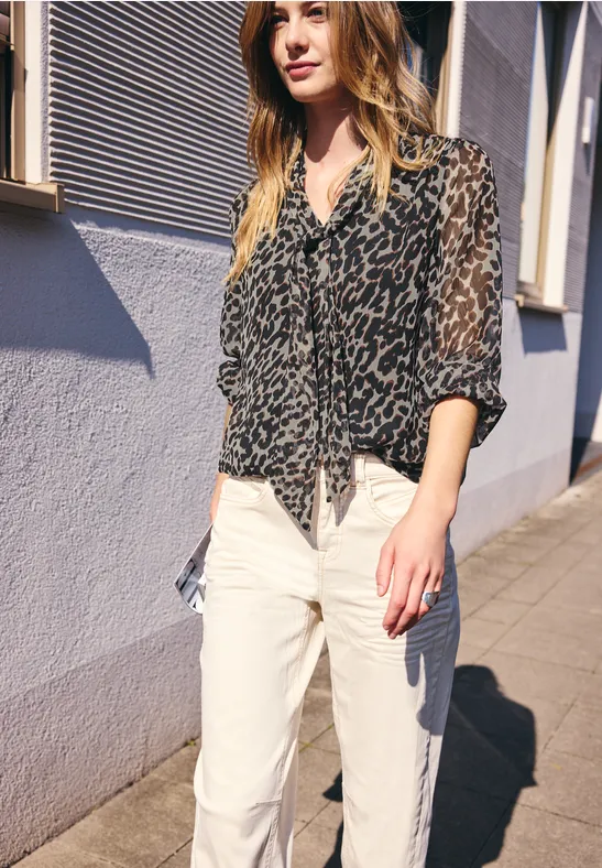 Femme en blouse léopard transparente noire et jean blanc, marchant au soleil le long d’un mur de maison.