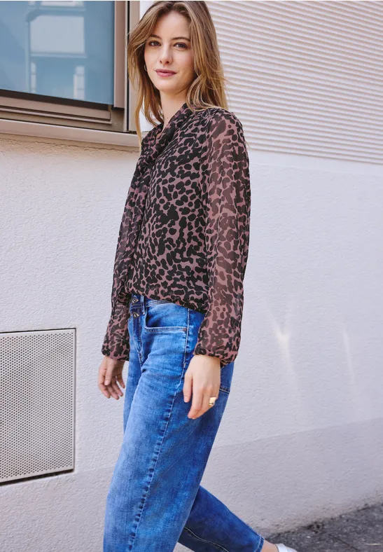 Jonge vrouw leunt nonchalant tegen een lichte muur, draagt een transparante luipaardblouse en blauwe jeans.