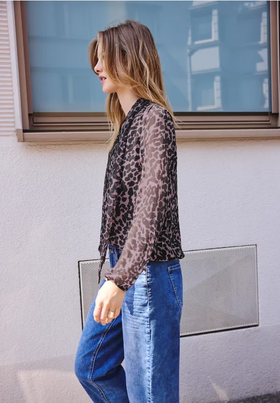 Een vrouw op de zijkant draagt een zwarte, gebloemde blouse en blauwe jeans voor een muur.