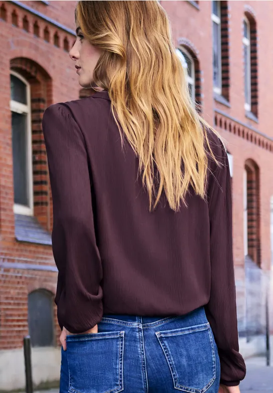 Vrouw van achteren in bordeaux blouse en spijkerbroek, voor een gevel van rode baksteen.