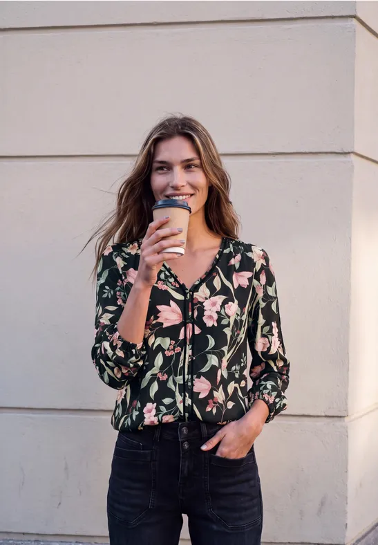 Eine lächelnde Frau mit floralem Oberteil und Jeans hält einen Kaffeebecher vor einer Wand.