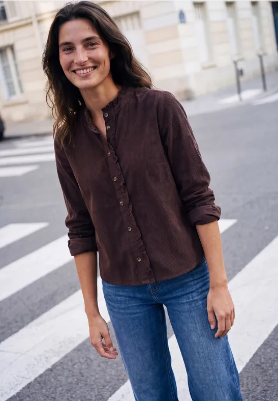 Auf einer Straße steht eine lächelnde Person mit braunem Hemd und Jeans.