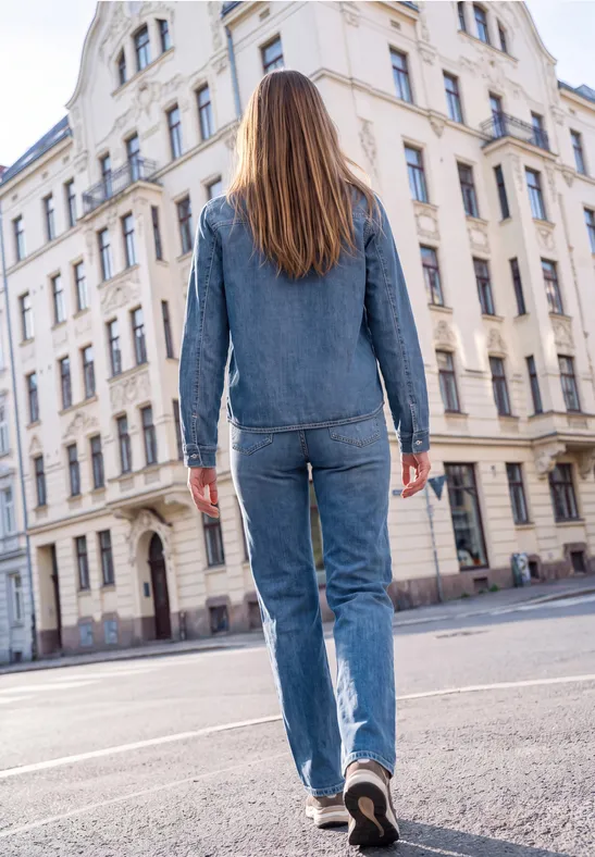 Eine Frau in Jeans-Outfit läuft auf einer Stadtstraße an einem alten Gebäude vorbei.