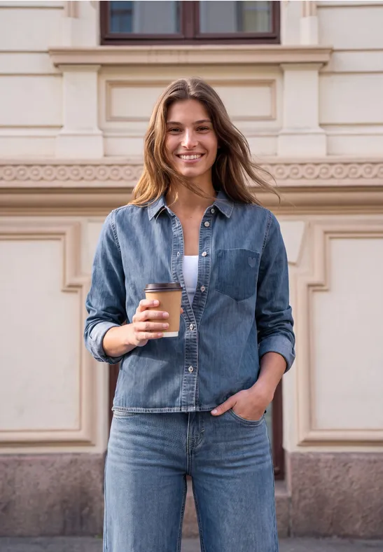 Junge Frau steht draußen vor einem Gebäude, lächelt und hält einen Coffee-to-go in der Hand.