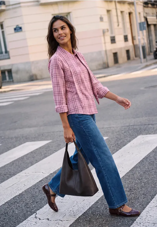 Frau in rosa Bluse und Jeans überquert lächelnd eine Straße, trägt eine Tasche in der Hand.