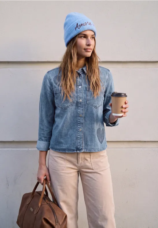 Frau in Jeanshemd, beiger Hose und blauer Mütze hält einen Kaffee und eine braune Tasche.