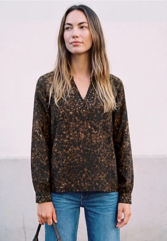 Frau in gemusterter dunkler Bluse und blauen Jeans vor hellem Hintergrund.