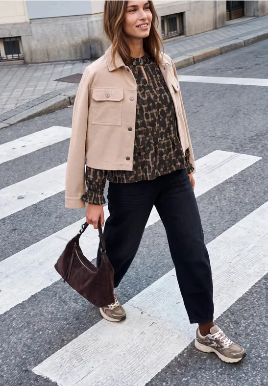 Eine Frau überquert einen Zebrastreifen in lässigem Outfit: helle Jacke, gemusterte Bluse, schwarze Hose und Sneaker.