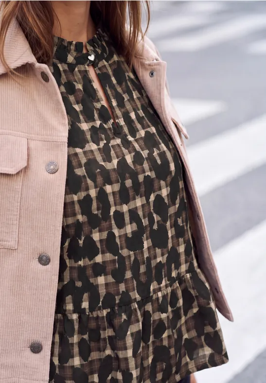 Eine Frau trägt eine rosa Jacke über einer dunkelgrünen gemusterten Bluse.