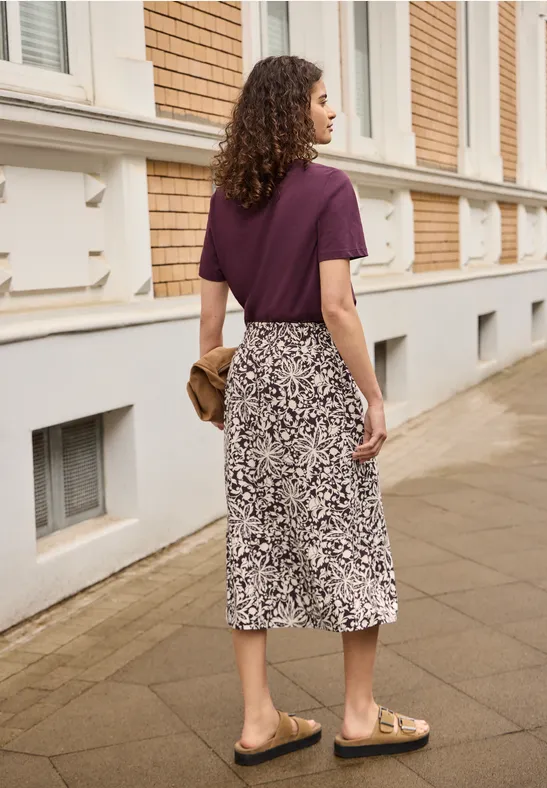 Vrouw van achter gezien in paars T-shirt en gemengde midi-rok, met clutch en sandalen op een stoep.