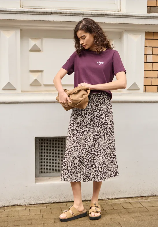 Vrouw in paars T-shirt, midirok met print en sandalen, met tas voor een gevel.