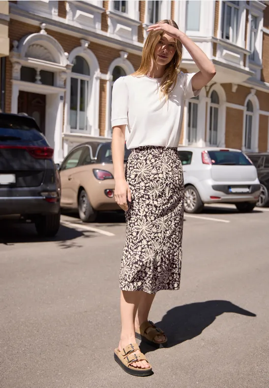 Frau in weißem T-Shirt und gemusterter Midirock mit Sandalen steht sonnig auf der Straße.