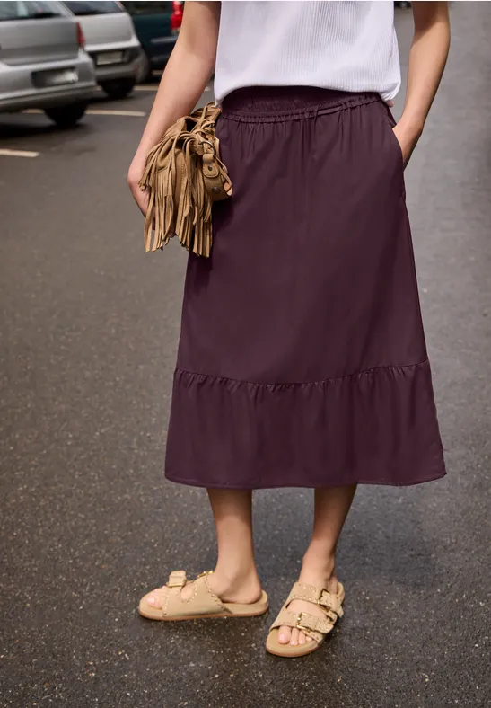 Vrouw met wit shirt en pruimkleurige midirok, draagt sandalen en een bruine franjehandtas op nat asfalt.
