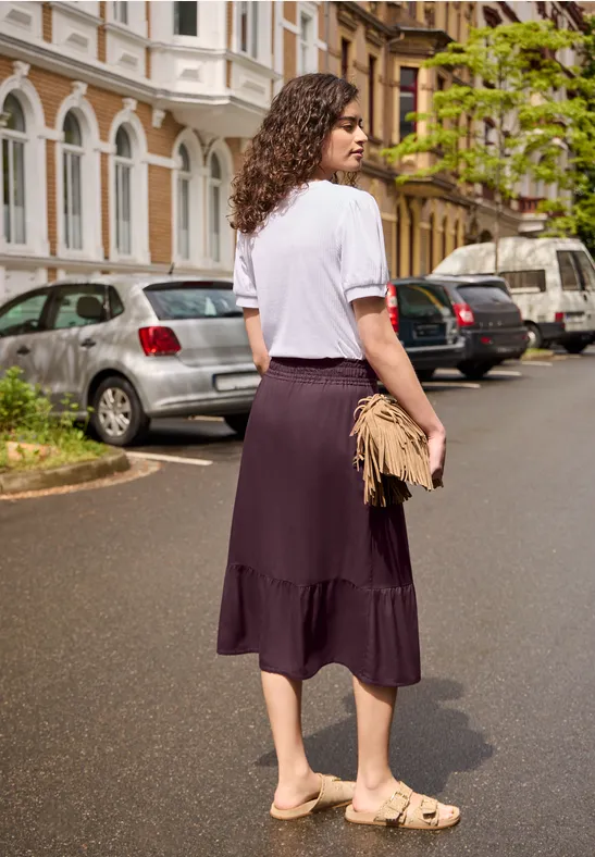 Een vrouw met krullend haar, wit T-shirt en paarse rok, met franjes-tas en sandalen, wandelt over straat.