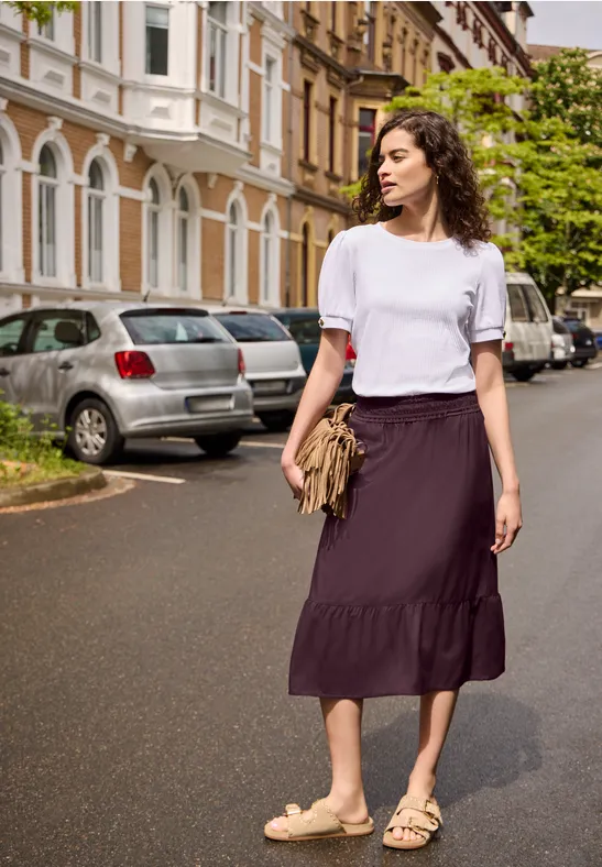Een vrouw in een wit T-shirt, paarse rok en sandalen wandelt over een woonstraat met geparkeerde auto's.