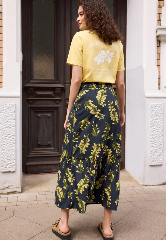 Eine Frau in gelbem T-Shirt und dunkelblauem Maxirock mit gelbem Blumenprint steht vor einem Hauseingang.