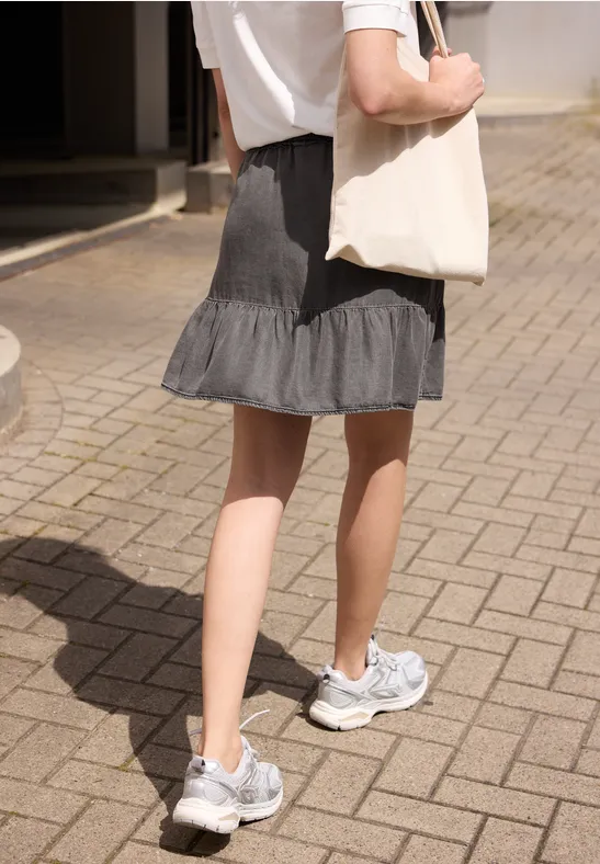 Frau von hinten in weißem Shirt, grauem Rock, Sneakern trägt weiße Tasche auf gepflastertem Weg.
