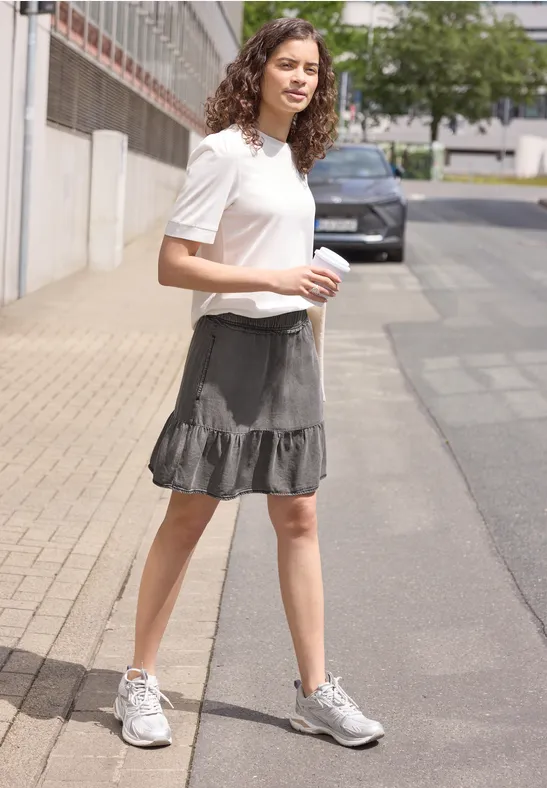Eine junge Frau geht auf dem Gehweg, trägt weißes T-Shirt, grauen Rüschensaumrock und weiße Sneaker.