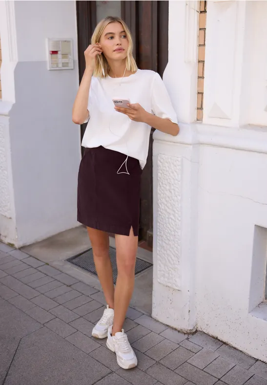 Jongedame in wit t-shirt, donkere rok en sneakers, staand op de stoep met smartphone in de hand, kijkt opzij.