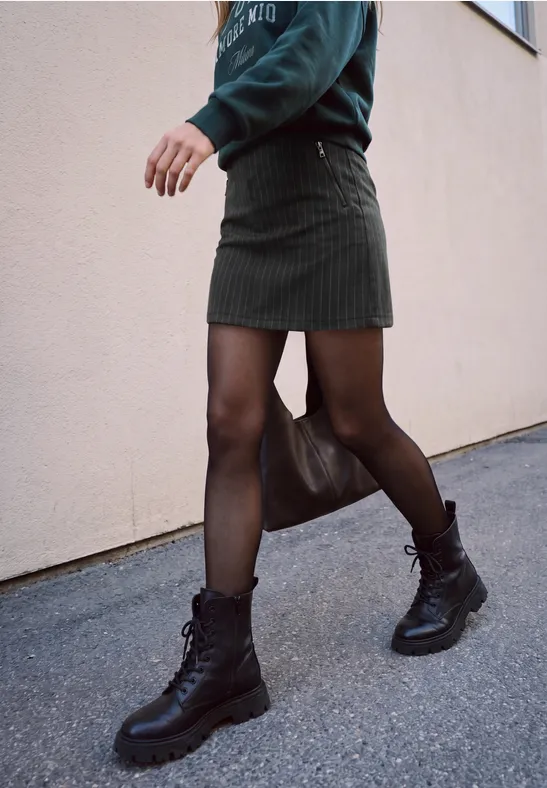 Eine Person in schwarzer Strumpfhose, kurzem Rock und Boots läuft an einer Hauswand entlang.