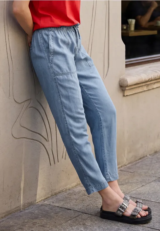 Person lehnt an Wand, trägt hellblaue Bundfalten-Hose, rotes Top und schwarze Sandalen mit Schnallen.