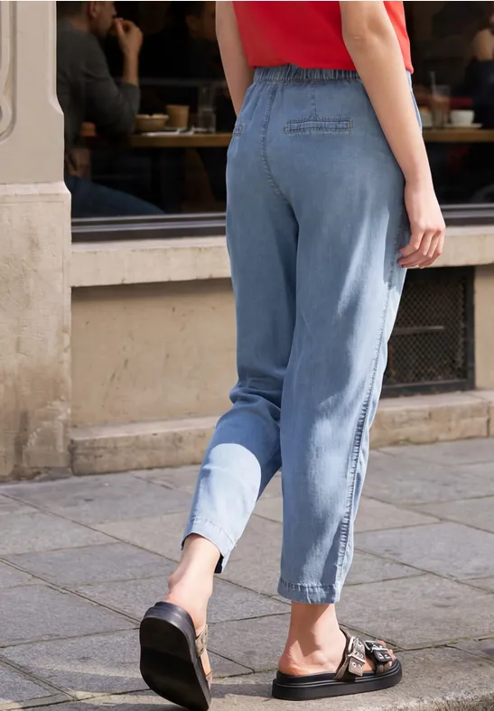 Eine Person in Jeanshose und Sandalen steht vor einem Café auf dem Gehweg.