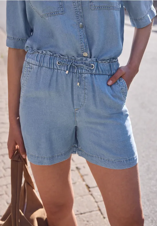Vrouw in een denim playsuit met elastische taille en zakken, handen in de zakken.