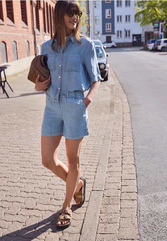 Een vrouw in jeansjasje en korte broek, met clutch in de hand en sandalen, loopt over een zonnig trottoir.