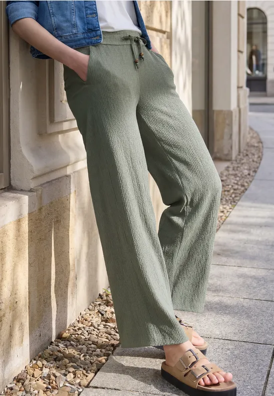 Personne en pantalon large en lin olive et sandales beiges appuyée contre un mur en pierre dehors.