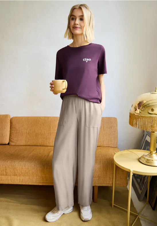Person in bordeauxrotem T-Shirt mit Schriftzug hält Tasse, vor gelbem Sofa und goldener Lampe.