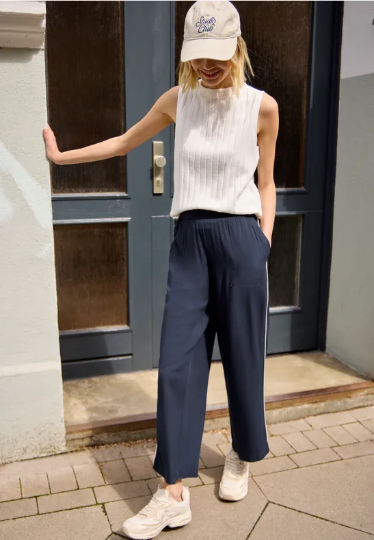 Frau in weißem ärmellosem Top, navy Culottes, weißen Sneakern und Baseballcap lehnt an Tür.