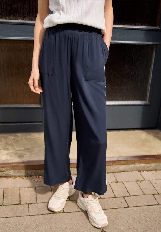 Person trägt weite dunkelblaue Hose, weiße Sneaker und helles Top, stehend vor Tür.