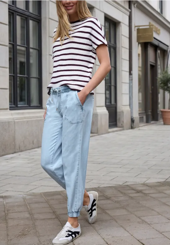 Vrouw in gestreept shirt en lichtblauwe jeans, hand in zak, nonchalant lopend over de stadstraat.