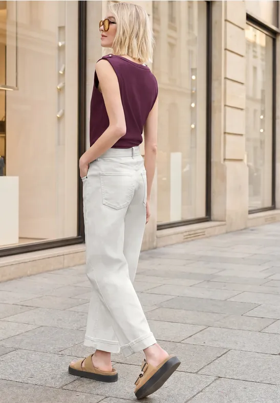 Eine blonde Frau in lila Top und weißen Jeans schlendert über eine Straße mit schicken Schaufenstern.