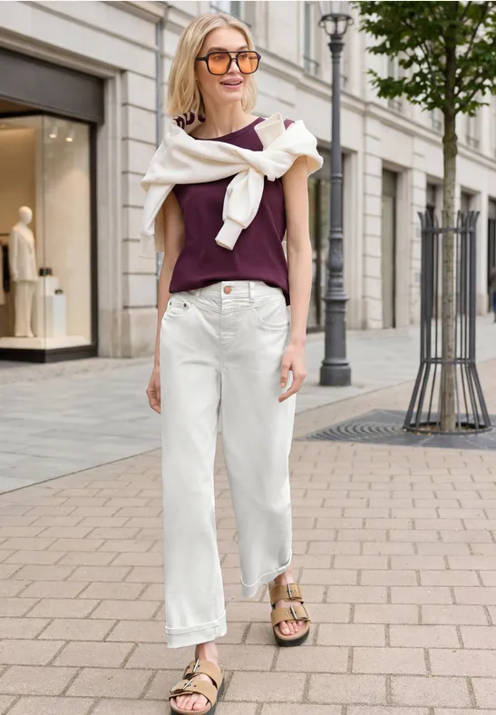 Frau in lila Top, weißer Hose und Sandalen schlendert lässig die Straße entlang.