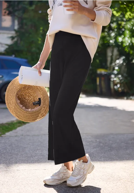 Eine Person in schwarzen Culottes, weißen Sneakern und mit Strohhut spaziert.