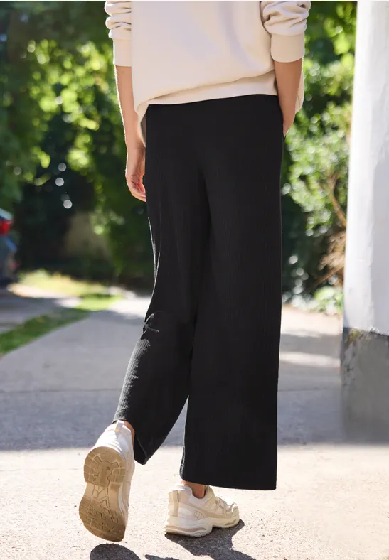 Rückansicht einer Person in schwarzen, weiten Hosen und weißen Sneakern, die auf einem sonnigen Gartenweg läuft.