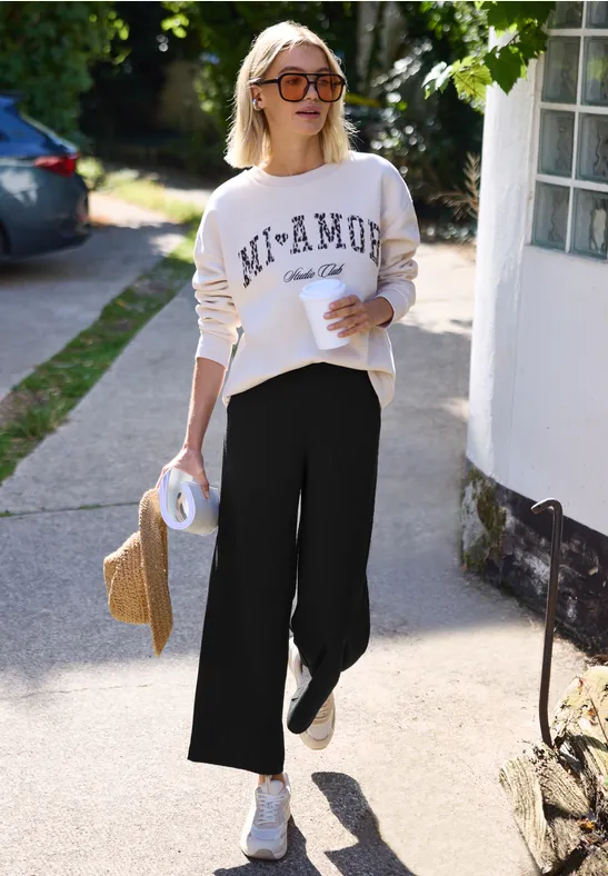 Frau mit Sonnenbrille, weißem Sweatshirt und Culotte spaziert entspannt einen Gehweg entlang und hält einen Strohhut.