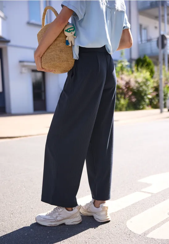 Person in weiten schwarzen Culottes, weißen Sneakern und Strohtasche auf sonnigem Gehweg.