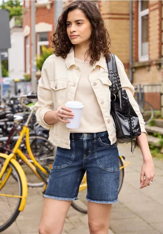 Frau spaziert mit Coffee-to-go in der Hand, trägt Jeans-Shorts und helle Jacke in einer Großstadt.