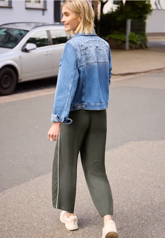 Person in blauer Jeansjacke und grünen Jogginghosen mit Seitenstreifen läuft auf der Straße.