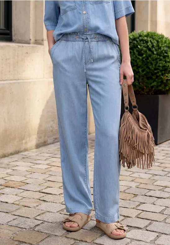 Person trägt hellblaues Denim-Top und weite Jeans, hält Fransen-Tasche, Sandalen.