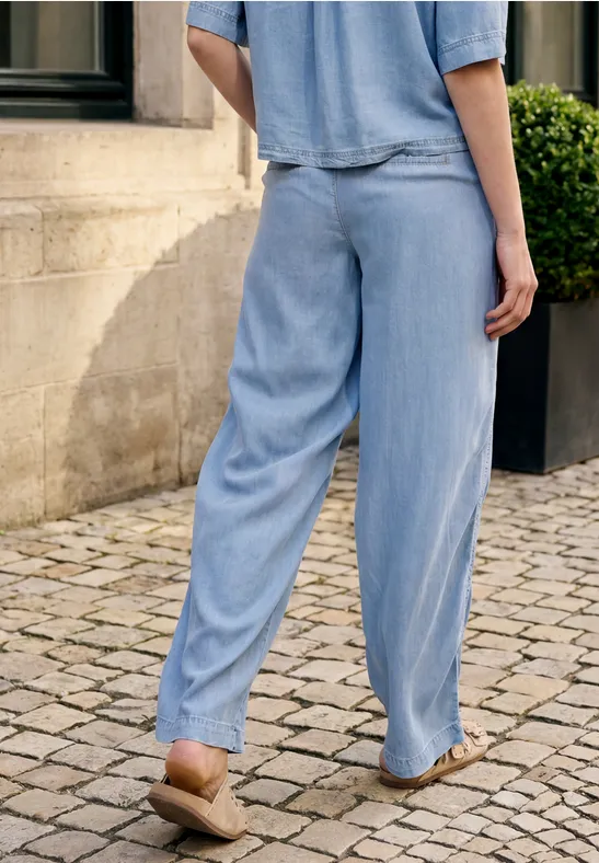 Eine Person trägt lockere hellblaue Hosen und Sandalen auf gepflastertem Boden vor einer Hauswand.