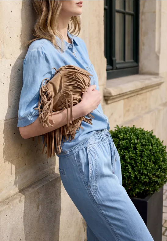 Frau in hellblauem Jumpsuit lehnt an Hauswand und hält eine braune Fransentasche neben einem Pflanzenkübel.