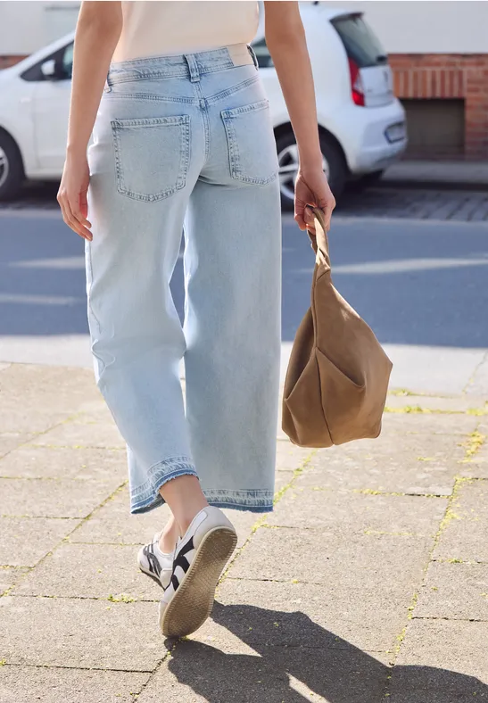 Persoon in lichtblauwe jeans en witte sneakers met een bruine tas loopt over het trottoir.