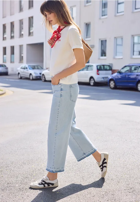 Een jonge vrouw, in spijkerbroek, T-shirt, rood sjaaltje en rugzak, loopt over straat.