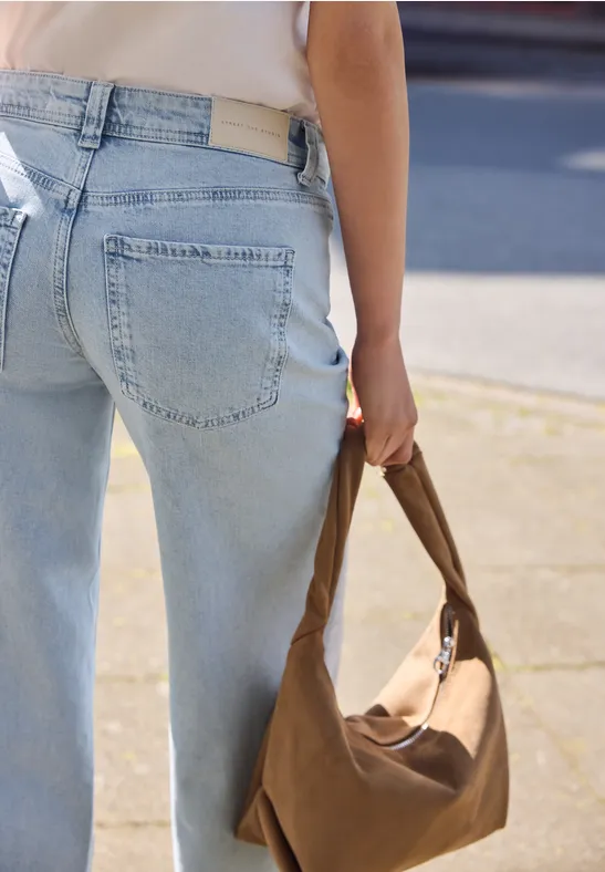 Persoon in lichtblauwe jeans draagt een bruine tas op een stoep gezien van achteren